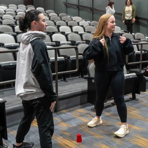 Two students stand opposite each other with a red cup on the ground in between them.