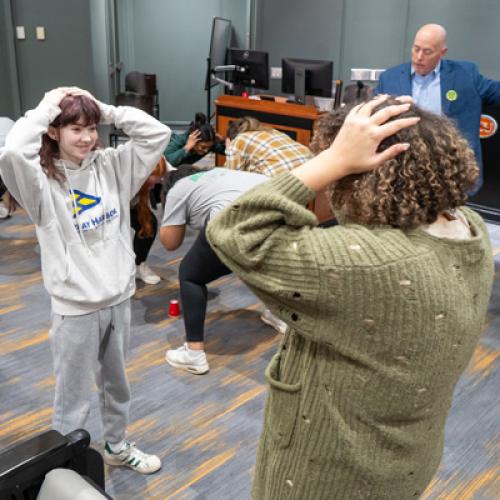 Students stand opposite each other with their hands on their heads, listening to Eric Smith explain the activity.