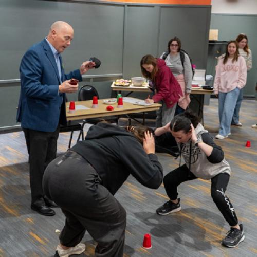 Two students stand opposite each other crouched over a red cup on the ground in between them with their hands on their heads listening to Eric Smith explain the activity.